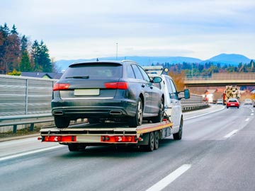 Ein Abschleppwagen von Unfallwagen Salzgitter transportiert ein schwarzes Auto auf einem flachen Anhänger entlang einer Autobahn, mit Bäumen und Bergen im Hintergrund.
