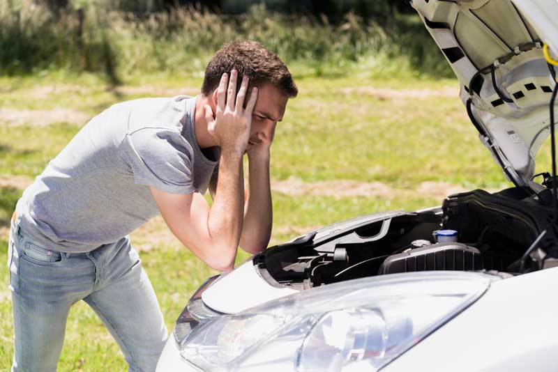 Autoankauf direkt in Ihrer Region – Fair und unkompliziert Ein Mann beugt sich über die offene Motorhaube eines Autos, sieht frustriert aus und ist auf einer Wiese an einem sonnigen Tag geparkt.